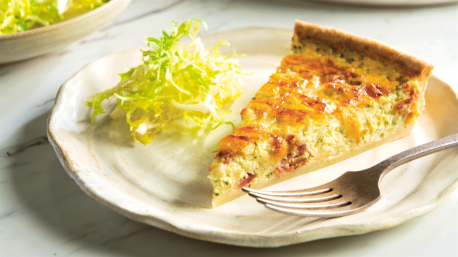 Slice of onion tart on a plate accompanied by a garnish of frisee and a fork